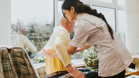 A young female carer helps an elderly male to get dressed by pulling on his jumper, as he sits in an armchair.