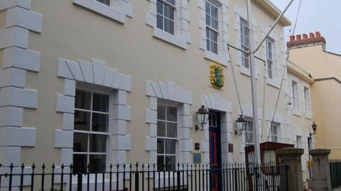 A yellow building with white around the windows. A crest sits above the front door while two lamps are either side of the door. A railing and two pillars sit in front of the building. Some of the neighbouring building including chimneys can be seen.