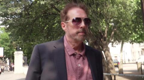 A man wearing a grey suit jacket and pink shirt walks along a tree-lined street. Only the top half of his torso and head are pictured. He has short brown hair and stubble and is wearing Aviator-style sun glasses.