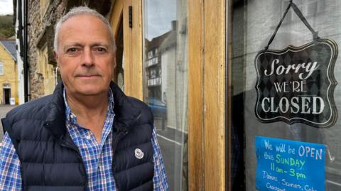 Steve Corrick, a grey haired man in a Moncler-branded functional gilet over a blue checked shirt. He stands outside his teashop in Axbridge town square. A sign on the door says "Sorry we are closed".