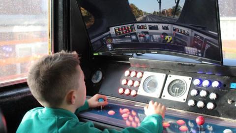 A pupil wearing a turquoise green jumper plays on a train simulator in the driver's cab of a Pacer train. 