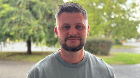 A man wearing a grey t-shirt stands outside. He is looking at the camera.