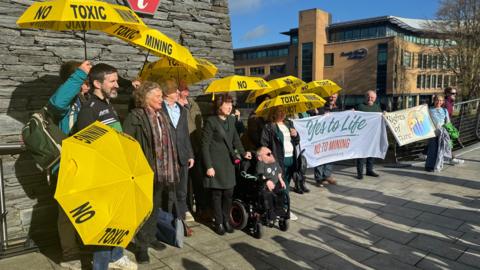 A number of people some holding yellow umbrellas saying no toxic mining stand outside a public building. Some of them are holding up a banner which says "Yes to life" in green text and "no to mining" in red text. Behind them a large brick and glass building can be seen.