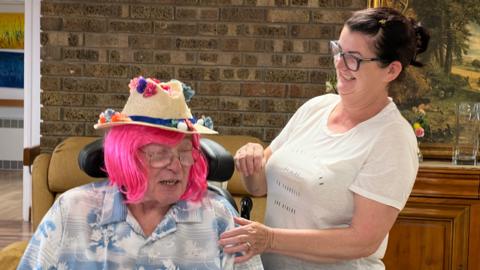 A man in a wheelchair wears a bright pink wig and a floral shirt and has a hat on with flowers attached to it while a woman smiles at him by his side