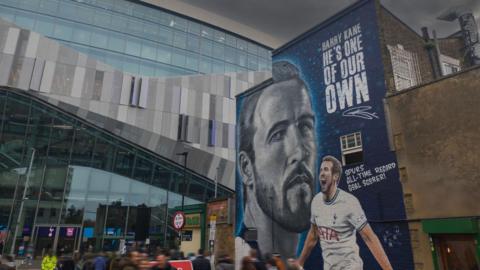Part of a large modern football stadium with an older brick building to the right of the image. There is a mural of footballer Harry Kane on the wall of the building. 