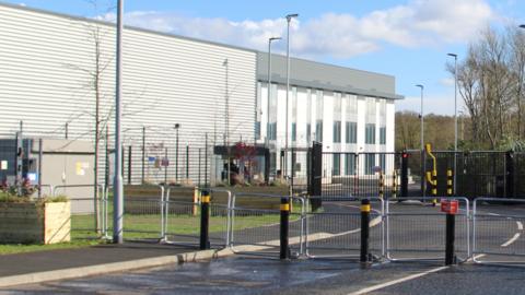 The exterior of a grey and white building with two rows of external temporary fencing and also black and yellow traffic blocking posts