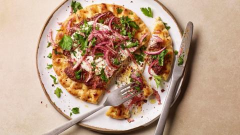 Flatbread topped with lamb, hummus, sliced red onion and parsley, on a white plate with a knife and fork