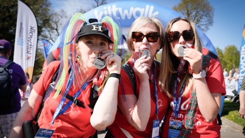Three kiltwalkers in red T-shirts, kissing their medals. They are wearing red T-shirts advertising a charity and Kiltwalk lanyards. The Kiltwalk finish line is behind them