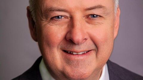 Ben Adams in an official portrait photo. He has blue eyes, grey hair and a smile. He is wearing a suit and against a plain grey background. 