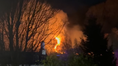 Orange flames and white smoke above the tree line covering most of the sky. There is a white chimney in front of the fire. The rest of the sky is dark.