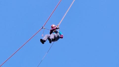 An elderly man abseils down a high building - all that is visible behind him is clear, blue sky. 