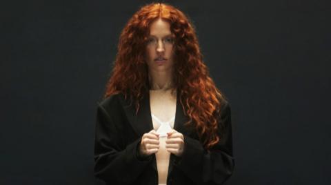 Jess Glynne, with long red hair, is looking at the camera. She is wearing a dark jacket and standing in front of a black background. 