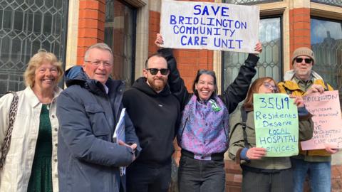 A group of six men and women in outerwear hold up placards with slogans reading: "Save Bridlington Community Care Unit".