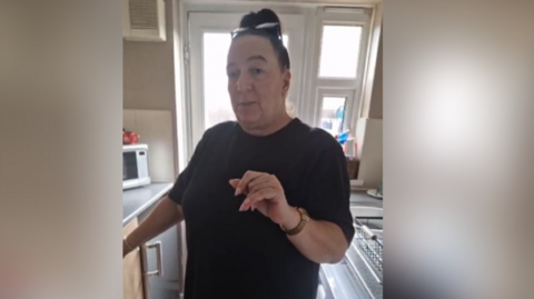 Claire has dark brown hair tied back with glasses on her head and is stood in her kitchen making a video. She is wearing a dark coloured top and had kitchen units behind her.