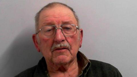 A police mugshot of a man in his early eighties. He has very short grey hair, a ruddy complexion, glasses and a short moustache.