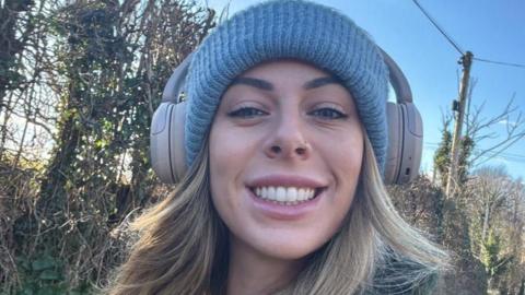 A woman smiles into the camera close up. She has long dark blonde hair under a light blue woolly hat. She is wearing headphones on top of the hat. Behind her are trees, a telegraph pole and blue sky.