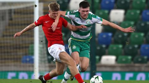 Two football players holding each other back as they run towards the ball. The male player on the left is a player for Larne FC, he wears a red top with white shorts, red socks and black football boots. His foot is closer to the white and green patterned ball. The male player on the right is a Shamrock Rovers player and he wears a green and white striped top, shorts and socks.