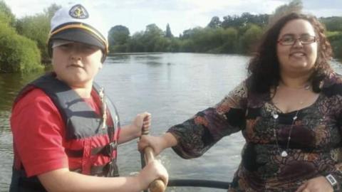 Tomas Cozens as a young boy with his mum Jo. They are on a boat on a river and are holding a wooden handle that steers the boat. Tomas is wearing a red t-shirt and a black and red life jacket with a white, Navy-style captain's hat. His mum has curly dark hair and rectangle-shaped glasses with a dark, patterned long-sleeved top on. The river and trees are visible in the background 