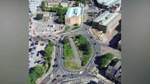 An aerial view of a roundabout with cars driving round it. The roofs of buildings and a car park dot the landscape along with a few with areas of trees and grass.