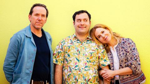 A family of three. One man stands in the middle, a woman hugs onto his left arm and leans her head on his shoulder. Another man stands to his right, arms folded behind his back. He looks less pleased than the other two