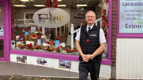A man with short grey hair standing in front of a funeral director's premises. The building's window frames are painted purple, with a seasonal window display featuring poppies and autumn leaves.