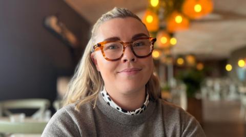 A woman with mid length blond hair and blue eyes. She is wearing thick tortoise shell framed glasses and smiling at the camera. She is wearing a stone grey jumper with a polka dot collar from her shirt visible.