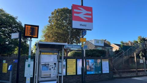A red railway sign which says Ifield on it. There is a self service ticket machine and the railway behind at ifield station