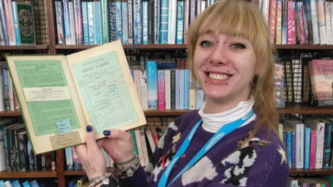 A woman with light hair wearing a purple jumper smiles and holds up an open copy of an old library book. An old-fashioned issue label is stuck inside the front cover.