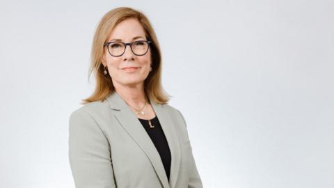 A woman wearing glasses is smiling at the camera. She is posing for a professional headshot. She has shoulder length blonde hair, a grey blazer and black top.