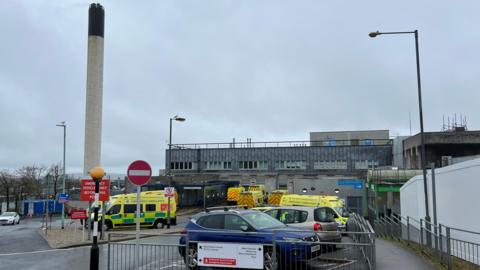 Several ambulances and a couple of cars parked outside Derriford Hospital in Plymouth on a gloomy day. A large chimney is in the background.