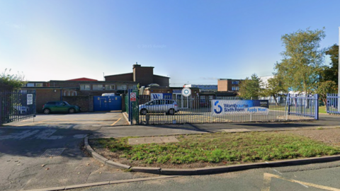 Bright blue sky can be seen with mainly brick, school buildings in the lower half of the image. A car park is surrounded by a fence which stands on the pavement with a grass verge on the other side. A banner advert for sixth form places is tied to the fence.