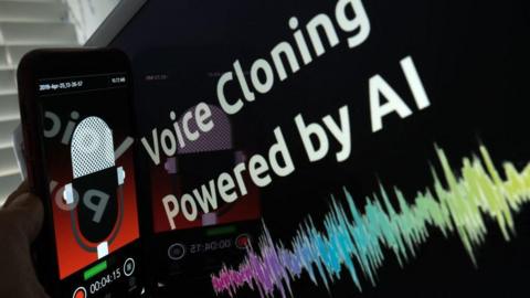 This picture is has a touchscreen mobile phone being held next to a screen which reads "voice cloning powered by AI". The mobile phone screen display has a animated radio microphone with a red background. The lower third of the mobile phone screen has a record button and a timing display. 
