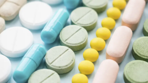 A stock image of lines of pills from above - from left, a line of round white pills, a line of pale blue capsules, a line of pale green round pills, a line of bright yellow small round pills, a line of pink capsules, then a line of pale green round pills