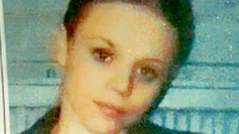 Donna Keogh with cropped dark hair and dark eyes. She is smiling with her head tilted to one side and standing in front of a light blue wall. The photo is very grainy.