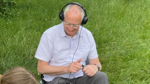 A man sat on a chair in the grass. He has on headphones which are connected to a small morse key in his hands with a black wire. He is wearing a light blue shirt with white stripes and grey canvas trousers. He is also wearing some clear rimmed glasses.