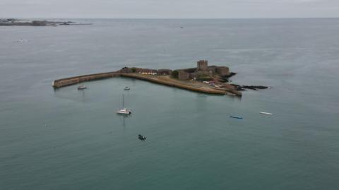 A drone shot of St Aubin's Fort. It is a concrete structure in the sea, with several small boats surrounding it.