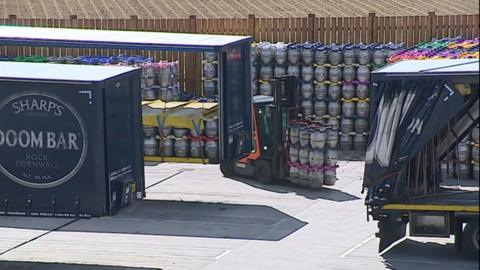 A forklift truck is carrying beer kegs across a tarmac area which has lorries and containers full of kegs. One of the containers is branded Sharp's Brewery Doom Bar. 
