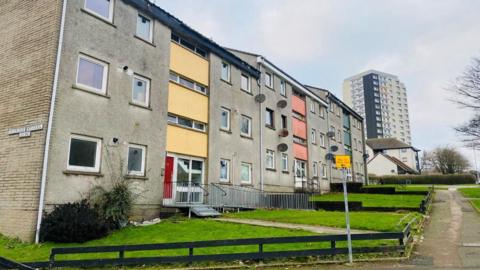 Blocks of flats, both three-storey and a high rise, with green gardens, next to a road, under a cloudy blue sky.