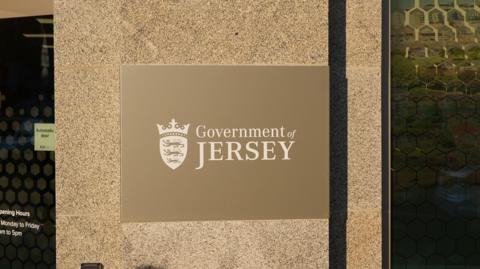 A gold and white coloured Government of Jersey sign on the side of a building. An automatic door is to the left of the sign.