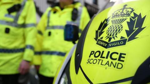 Police officers in yellow hi-vis jackets, next to a vehicle with a Police Scotland logo.