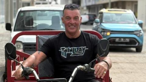 A man with grey hair and sunglasses on top of his head sits on a motorbike, holding the handlebars. He wears a black t-shirt and a bead bracelet. He looks to be on holiday.