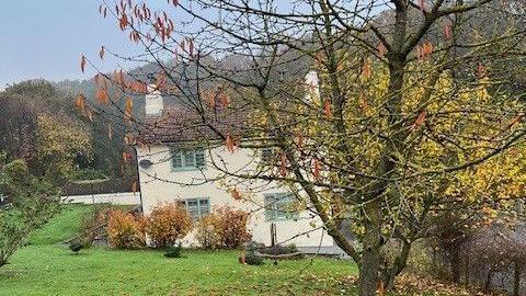 Hett Mill Cottage is a white two-storey house with pale blue window panes and a triangle roof. The property is in a secluded location, surrounded by lots of grassland and trees. 