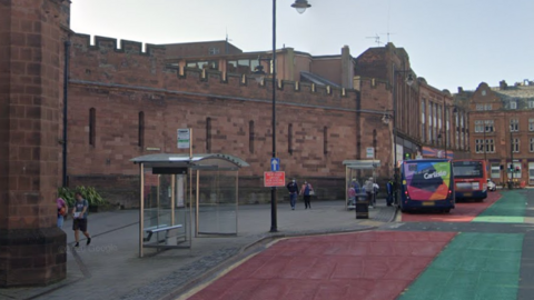 A Google Streetview screenshot of English Street. The road is flanked by the red-stone Citadel buildings on the left and shops on the right. There are two bus stops on the right and two buses, one stopped at the stop letting people on and off, and one further along at a light. The street has red markings where the buses should stop, next to green markings, on the tarmac.