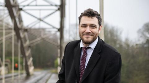 Hexham MP Joe Morris, wearing a black overcoat, shirt and tie and smiling with an out of focus bridge behind him.