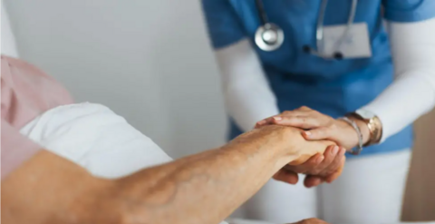 A nurse, wearing a blue top and white trousers, clasping the hand of a patient with both hands