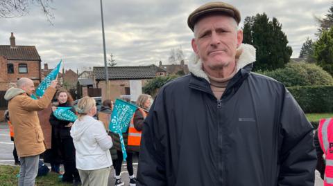 James Coulson is standing in a black coat and brown flat cap in front of a group of striking teachers. They have light blue flags showing they are from the National Education Union.