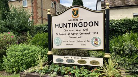 A sign in a planter which has lots of small plants in it. The sign says "Welcome to Huntingdon" in black writing. There are hedges and houses behind the sign.