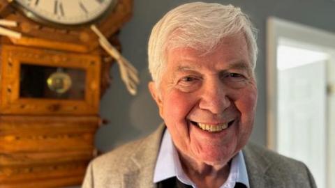 Nigel Thomas is smiling, standing beside a wall-mounted grandfather clock. He has white hair and is wearing a beige suit and pale blue, open-necked shirt.
