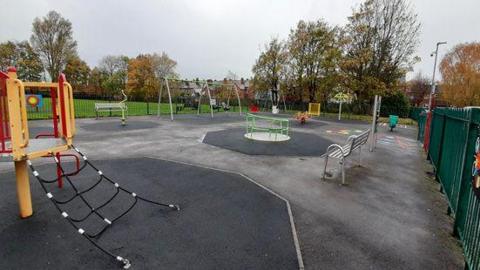 A play area, with colourful play equipment and green metal railign surrounding it. There are swings, a climbing frame and a wheelchair accessible roundabout.