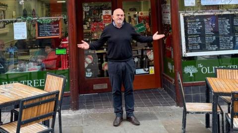 Kevin in front of his cafe. It has green signage and there are tables and chairs at the side of him. He has his hands up towards the sky.
